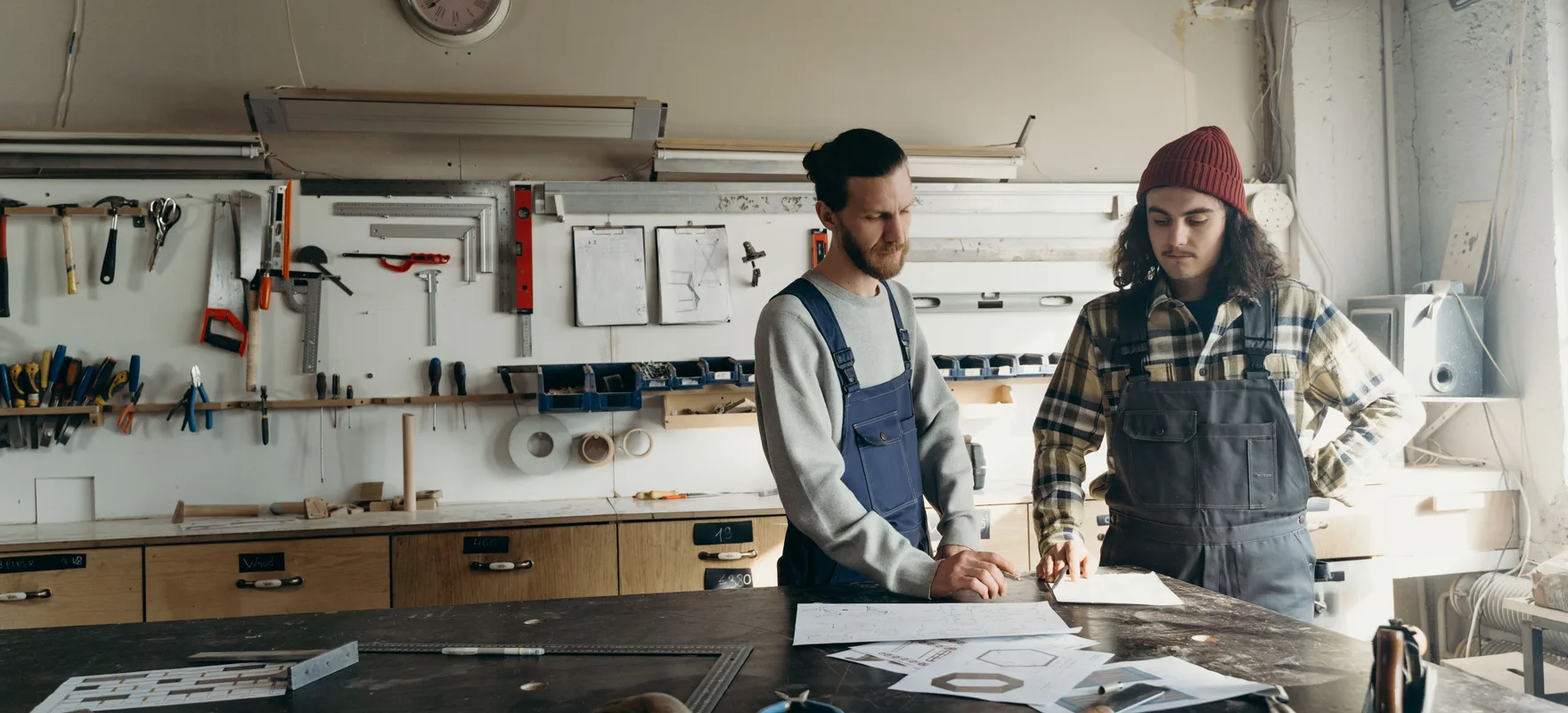 Zwei Handwerker stehen in einer Werkstatt an einer Werkbank und besprechen Zeichnungen, dahinter eine Wand mit Werkzeug.