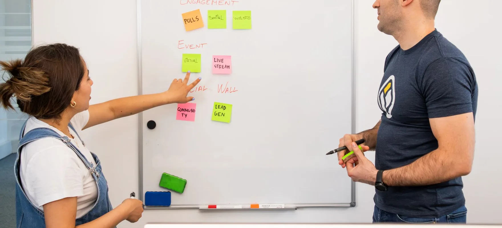 Eine Frau zeigt auf bunte Haftnotizen an einem Whiteboard, ihr gegenüber steht ein Kollege.
