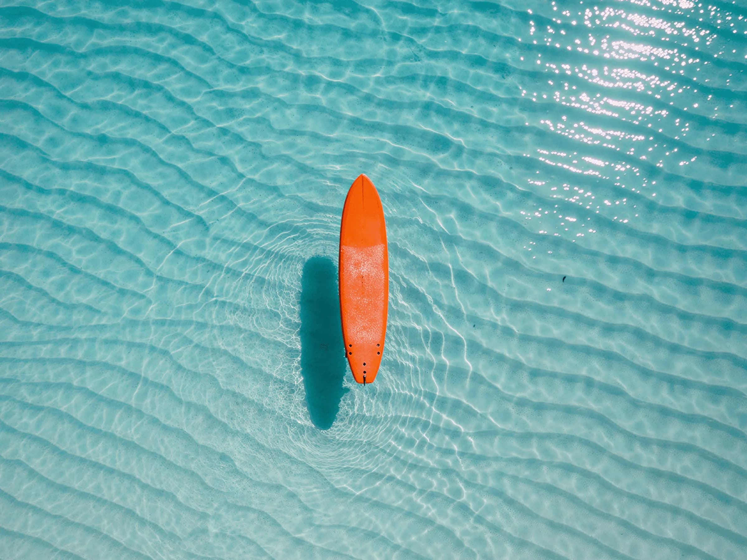 Blick von oben auf ein oranges Surfbrett, das allein auf flachem, klarem Wasser treibt