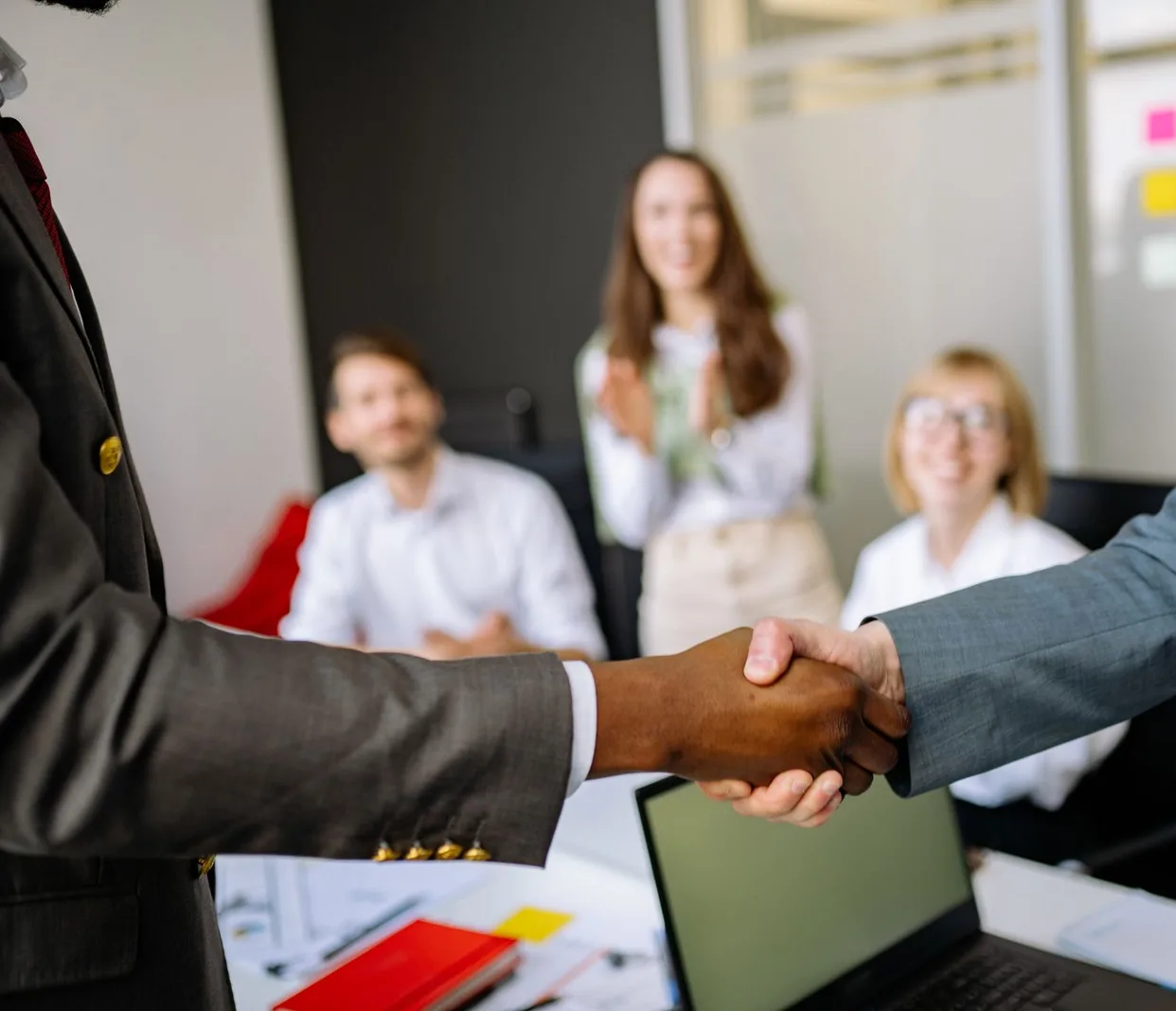 Zwei Menschen geben sich in einem Büro die Hand, im Hintergrund sitzt ein Team am Tisch.